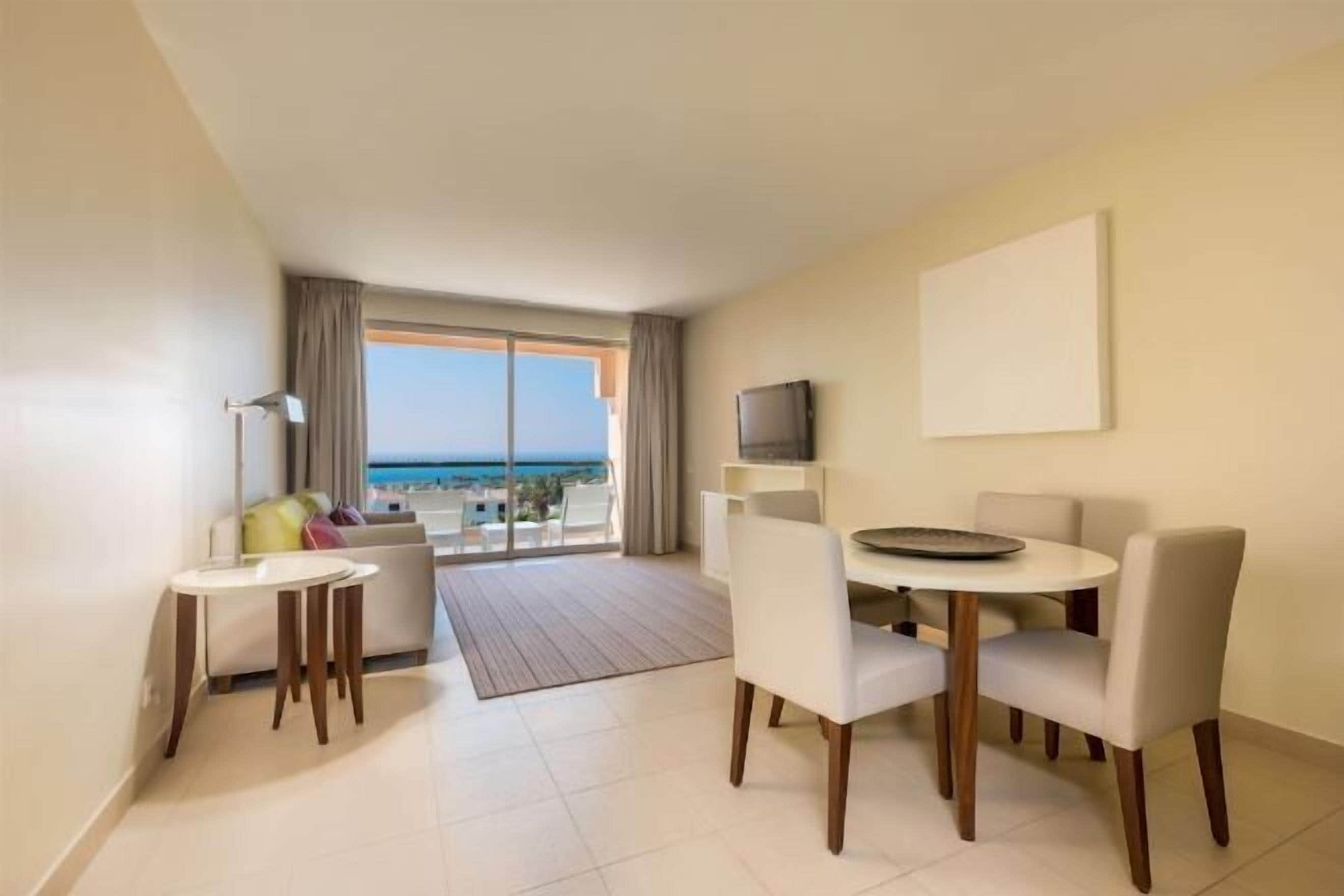 A modern hotel room with a balcony, a dining area with a round table and chairs, and a flat-screen TV mounted on the wall.