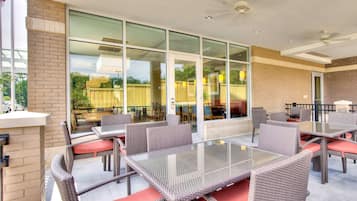 Un espace extérieur aménagé en terrasse, avec des tables à plateaux en verre et des chaises en osier, agrémenté d’un mur en briques et de grandes fenêtres.
