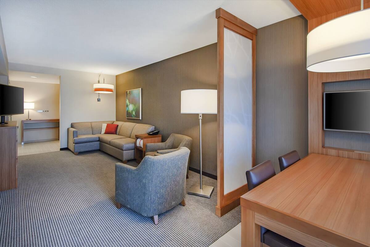 A hotel room with a sofa, armchair, dining table, TV, and lamp.