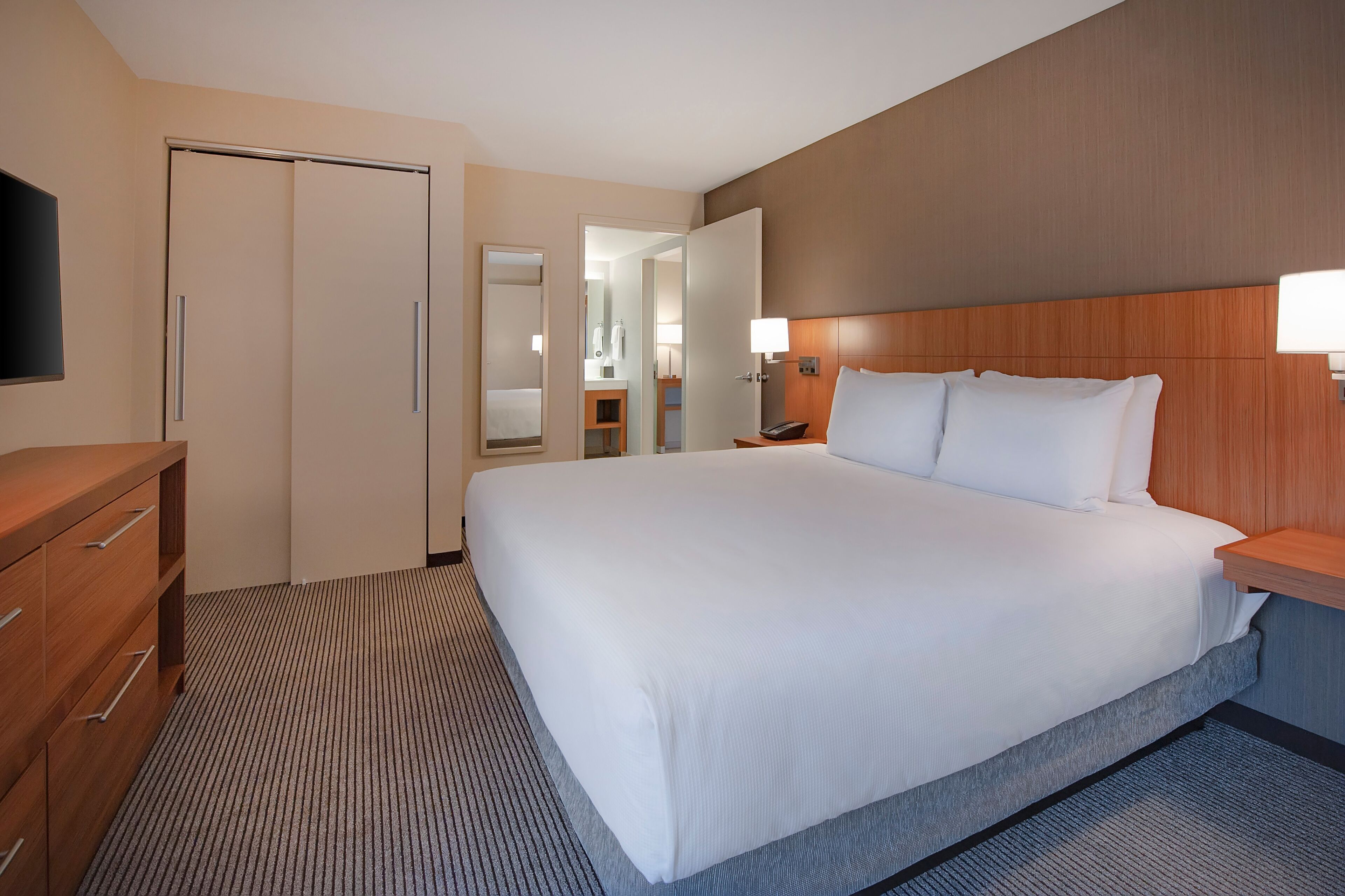 A hotel room with a large bed, a wooden dresser, a flat-screen TV, and a bathroom visible through an open door.