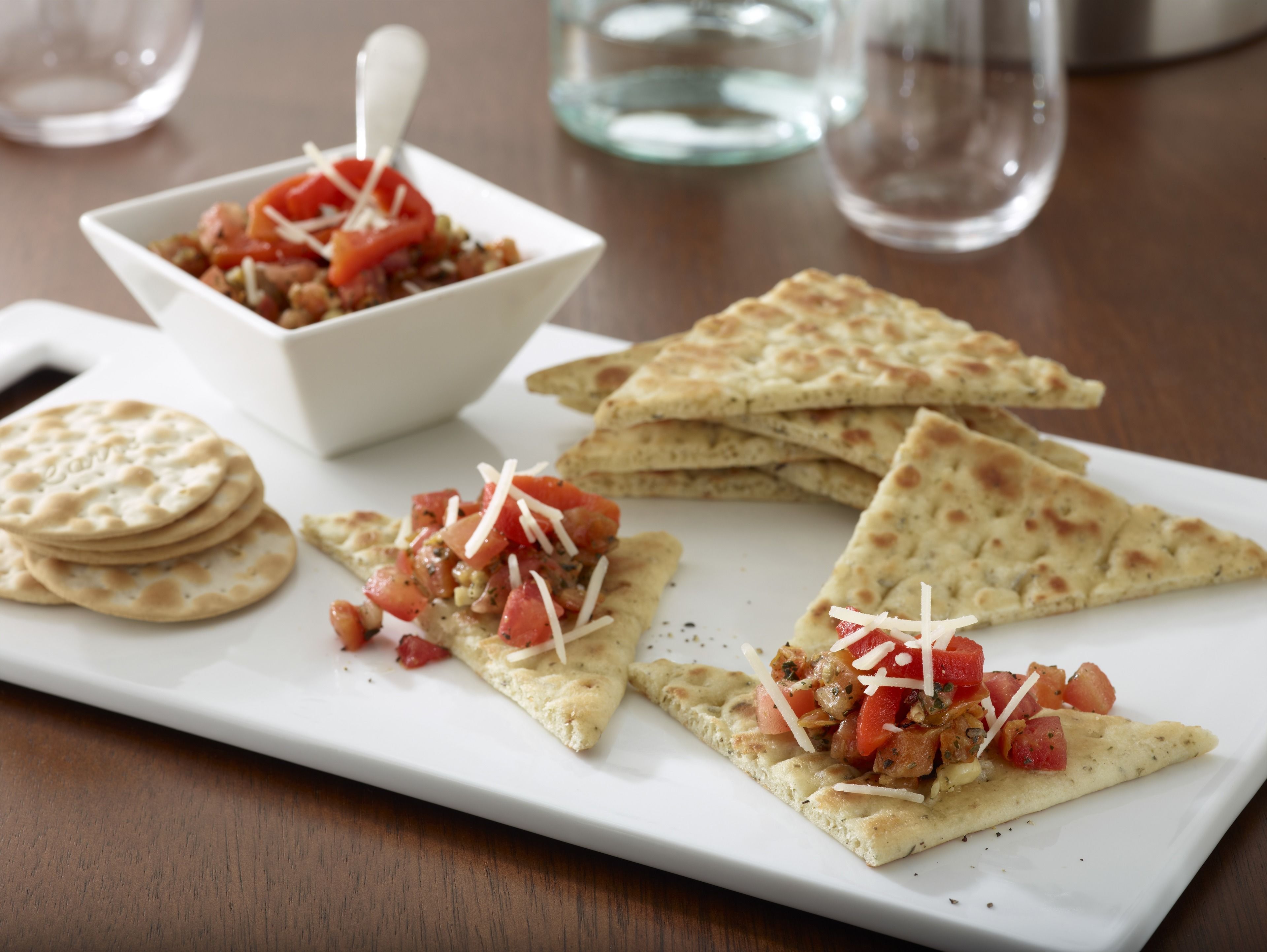 Un plato blanco rectangular con tres porciones triangulares de pan plano cubiertas de tomate y queso, un tazón pequeño de ensalada de tomate y una pila de panes planos redondos.