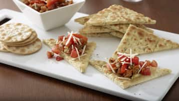 Un plato blanco rectangular con tres porciones triangulares de pan plano cubiertas de tomate y queso, un tazón pequeño de ensalada de tomate y una pila de panes planos redondos.