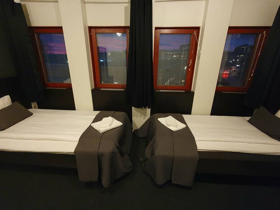 Una habitación con dos camas, cada una con colchón blanco y cabecera oscura. Hay dos ventanas con marcos de madera y persianas, y una cortina negra en el centro.