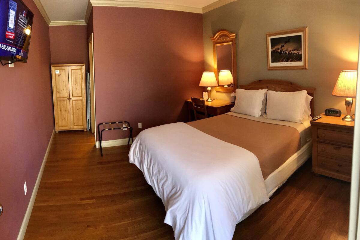 Ein Hotelzimmer mit einem großen Bett, Holzboden, einem Schreibtisch mit Stuhl, einem Nachttisch mit Lampe und einem gerahmten Bild an der Wand.