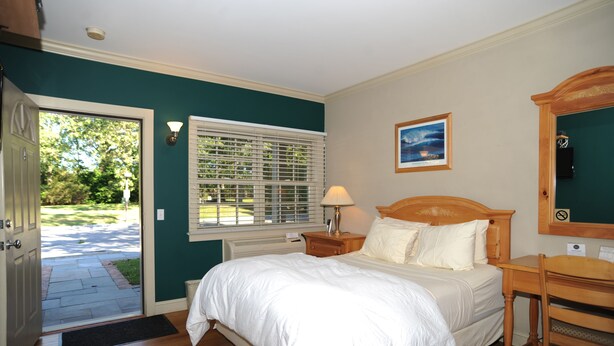 A bedroom with a bed, bedside table, lamp, and a view of the outdoors through a window. - Southampton Long Island Hotel (Southampton)