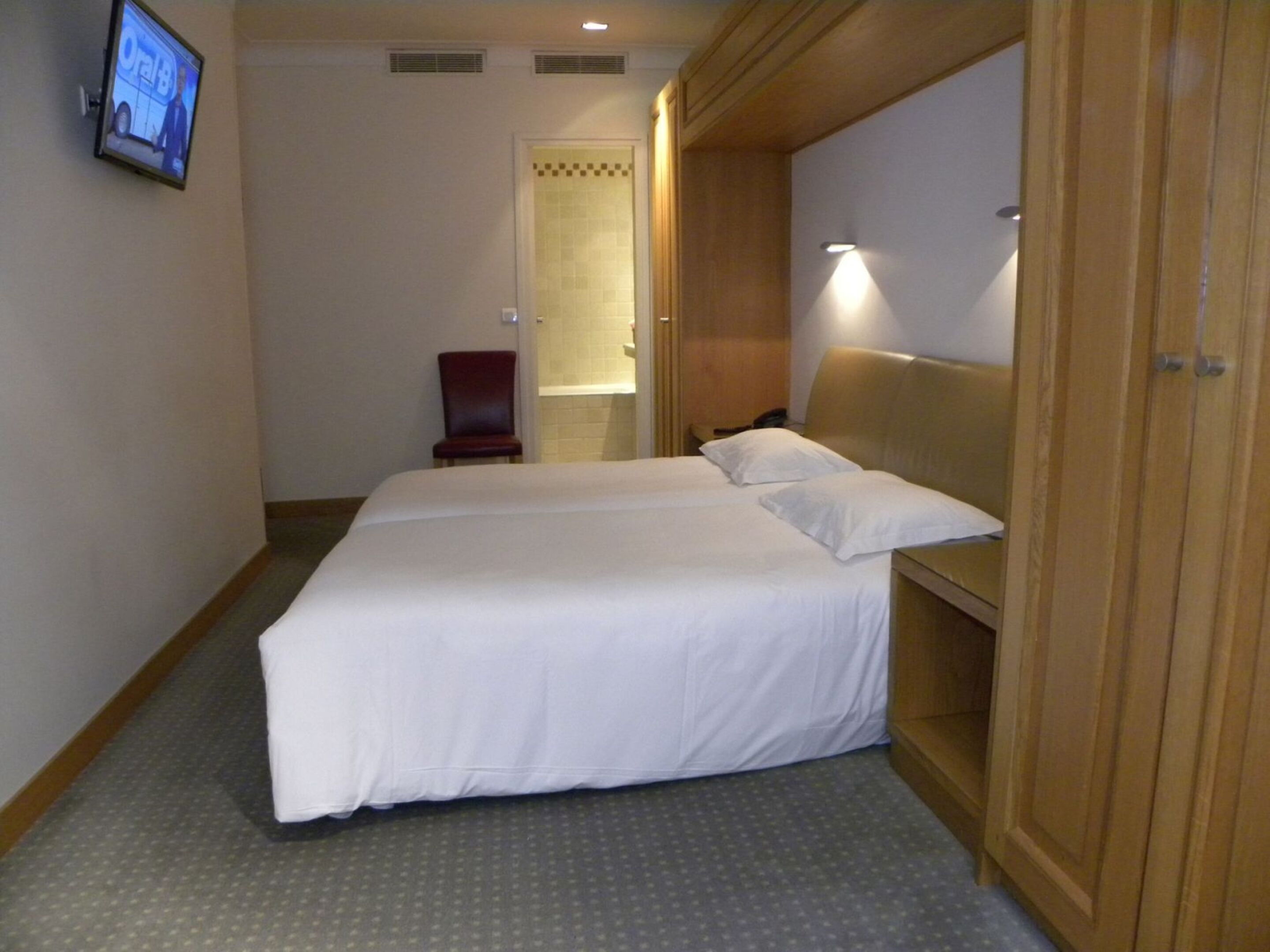 A hotel room with a large bed, a wooden wardrobe, a small chair, and a television mounted on the wall.
