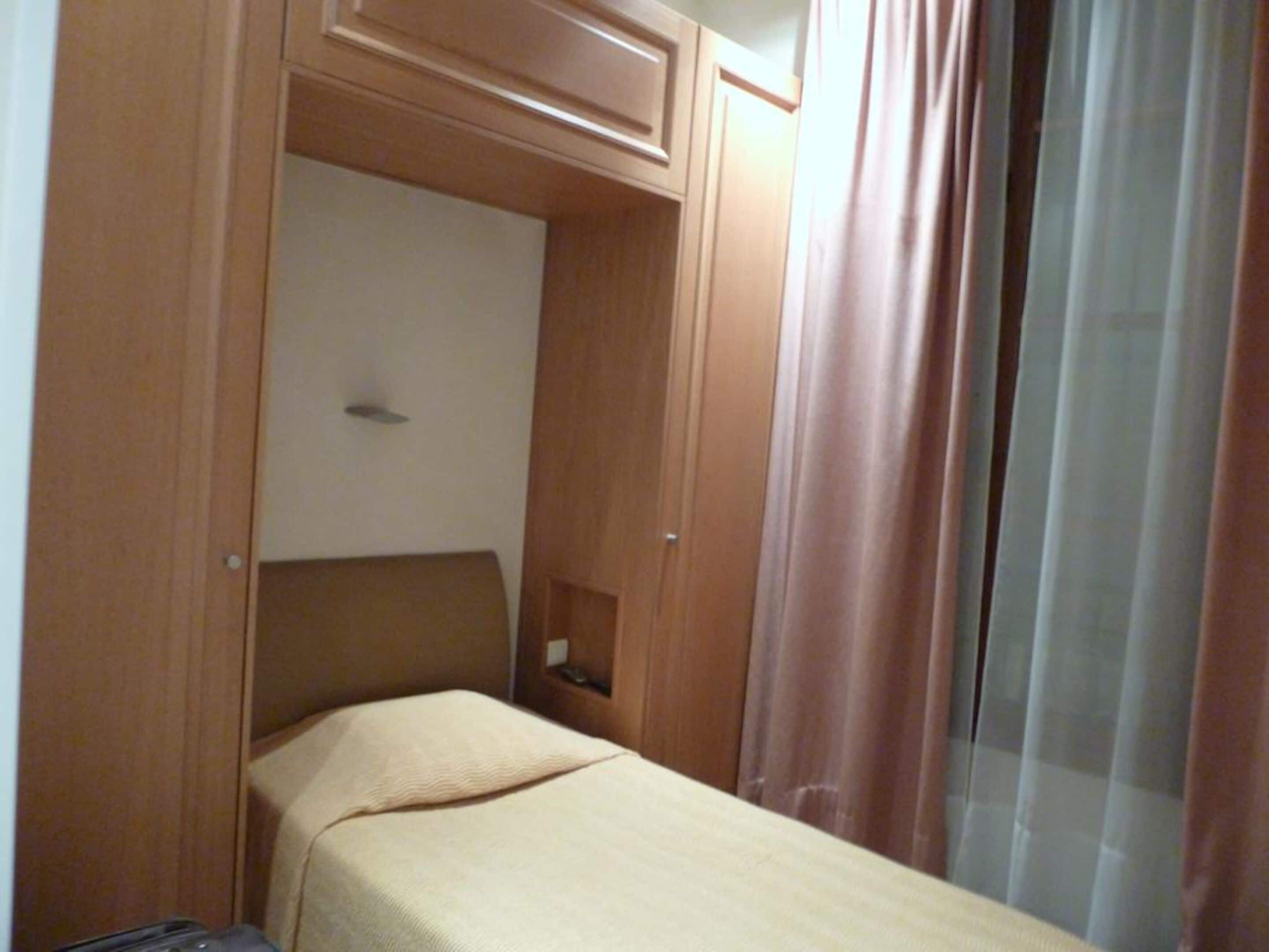 Habitación pequeña con una cama individual, cabecera de madera, mesita de noche y ventana con cortinas traslúcidas.