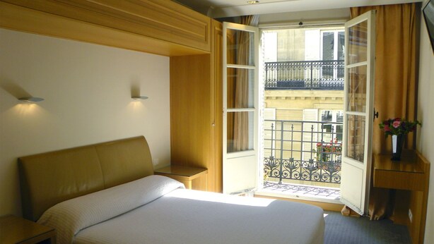 Habitación de hotel con cama, mesita de noche, escritorio con un florero y balcón con barandilla de hierro forjado y vistas a edificios.