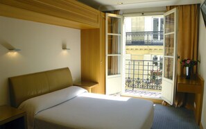 Habitación de hotel con cama, mesita de noche, escritorio con un florero y balcón con barandilla de hierro forjado y vistas a edificios.