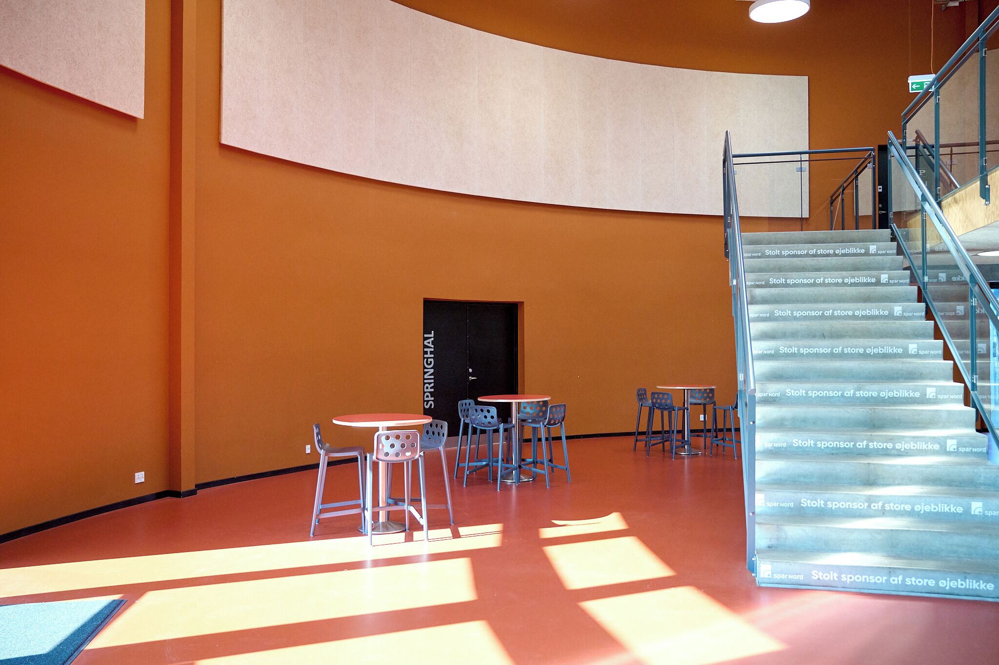 Un'ampia area interna con pareti arancioni, soffitto curvo e una scala con ringhiere in vetro. Tavoli e sedie disposti per sedersi.