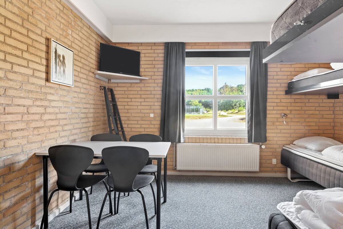 Una habitación con pared de ladrillo, ventana con cortinas, cama, mesa con sillas y televisor fijado en la pared.