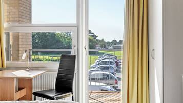 A hotel room with a bed, a desk, and a chair. There is a window with a view of parked cars and greenery outside.