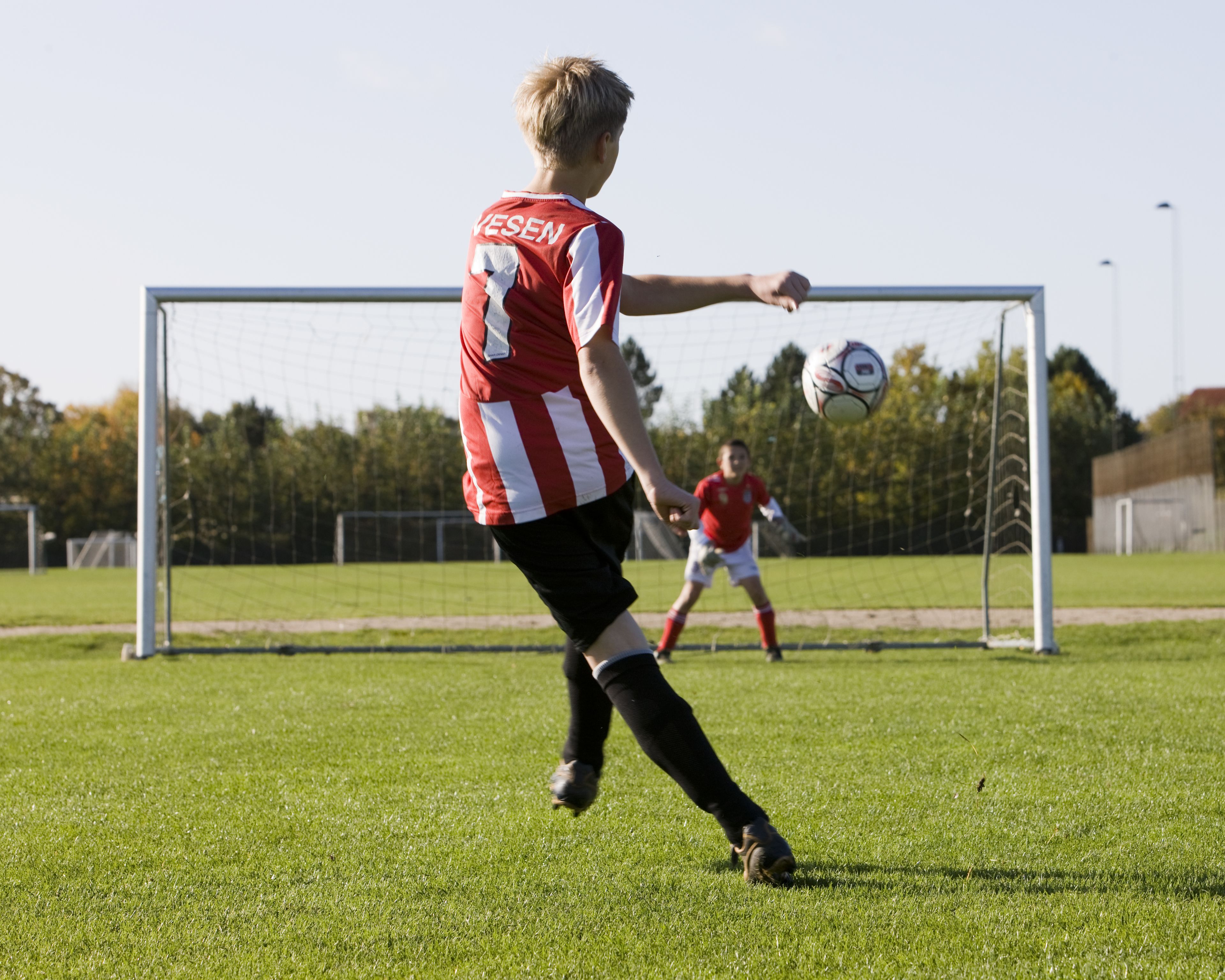 A soccer player in a red and white striped jersey with the name 'VESEN' on the back, preparing to kick a soccer ball towards a goal.