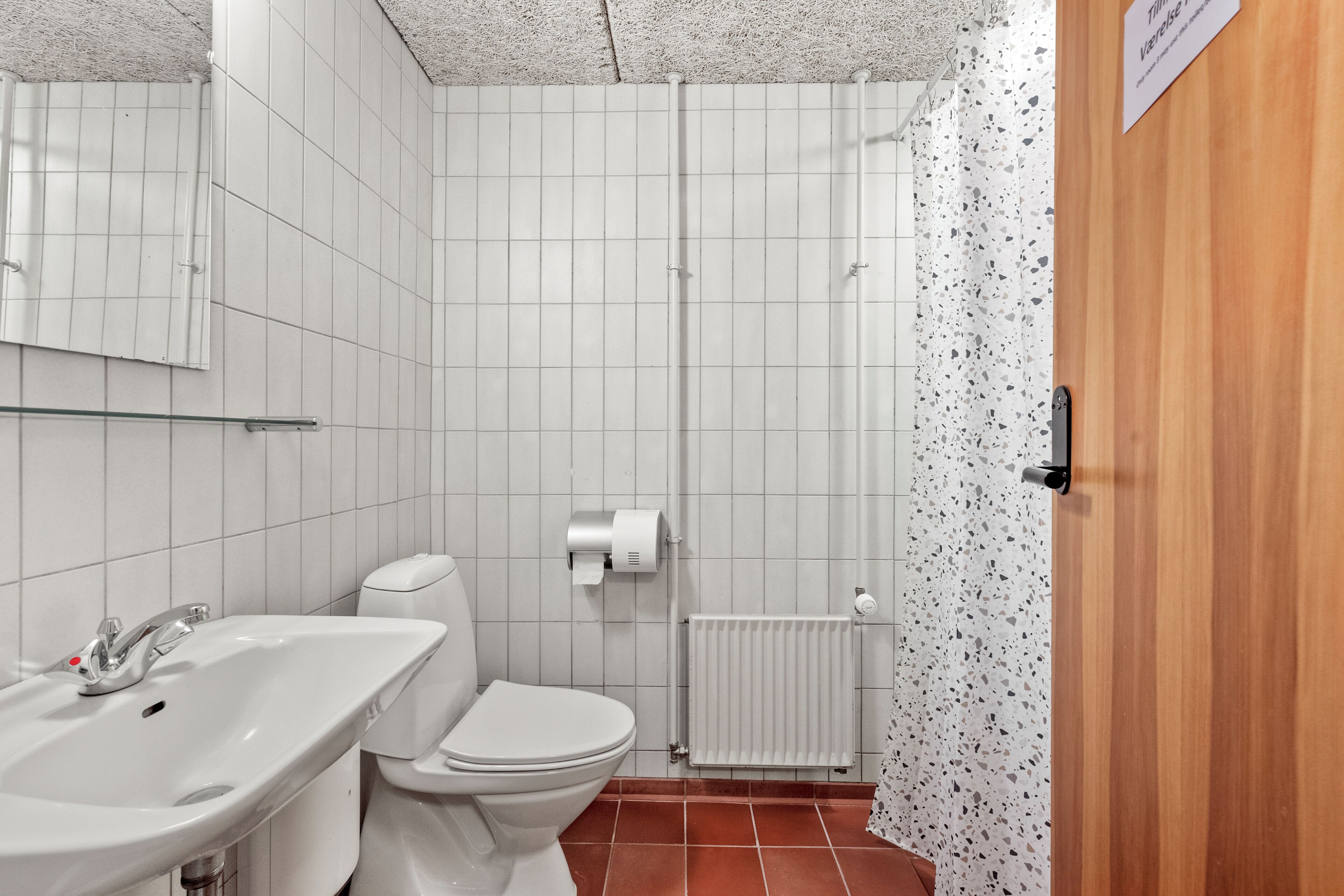 Un baño con azulejos blancos, un lavabo blanco, un inodoro, un dispensador de toallas de papel y un radiador.