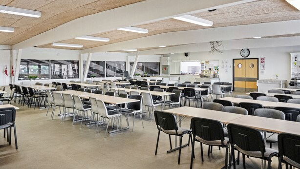 Una cafeteria espaiosa amb taules i cadires llargues, un rellotge a la paret i una finestra amb vista a l'exterior.