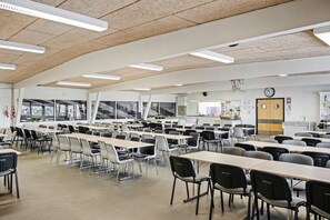 A spacious cafeteria with long tables and chairs, a clock on the wall, and a window with a view of the outside.