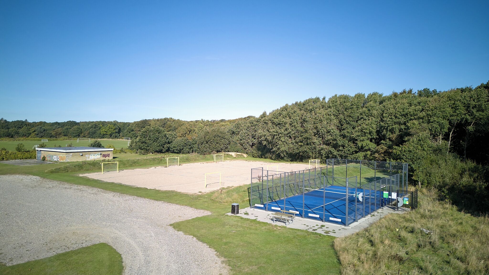 Instalación deportiva al aire libre con cancha de tenis, un edificio pequeño y un área boscosa en el fondo.
