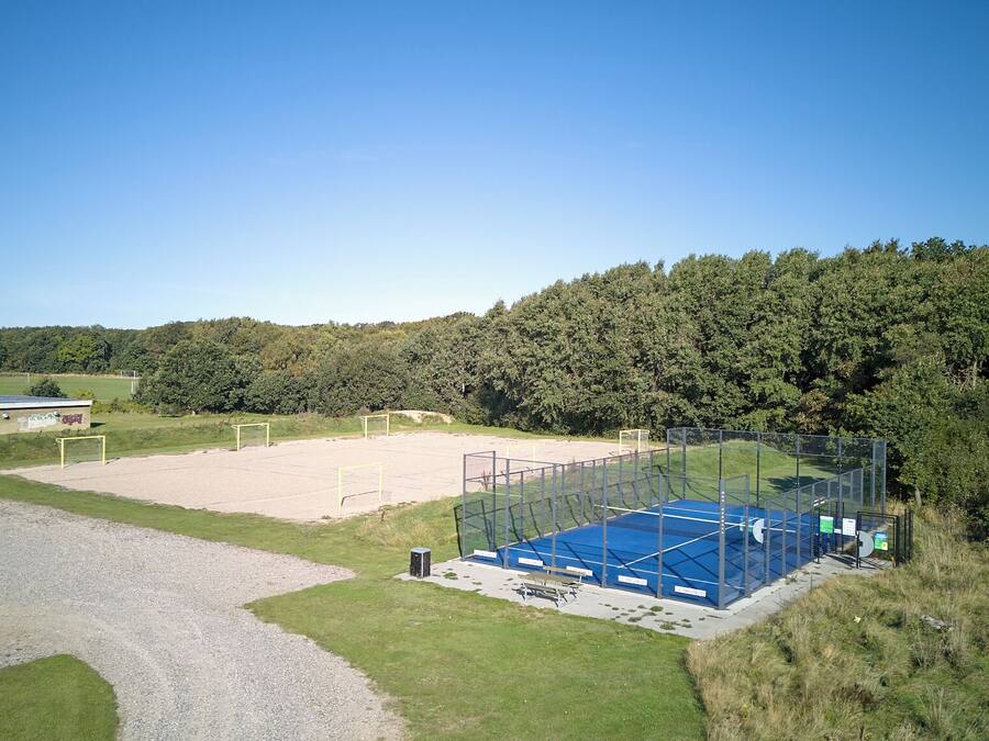Instalación deportiva al aire libre con cancha de tenis, un edificio pequeño y un área boscosa en el fondo.