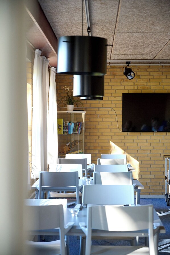 Un comedor con sillas y mesas blancas, una pared de ladrillo, un televisor montado y un accesorio de iluminación en el techo.