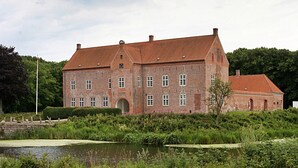 Un edifici gran de maó amb teulada vermella, envoltat de vegetació exuberant i un estany.