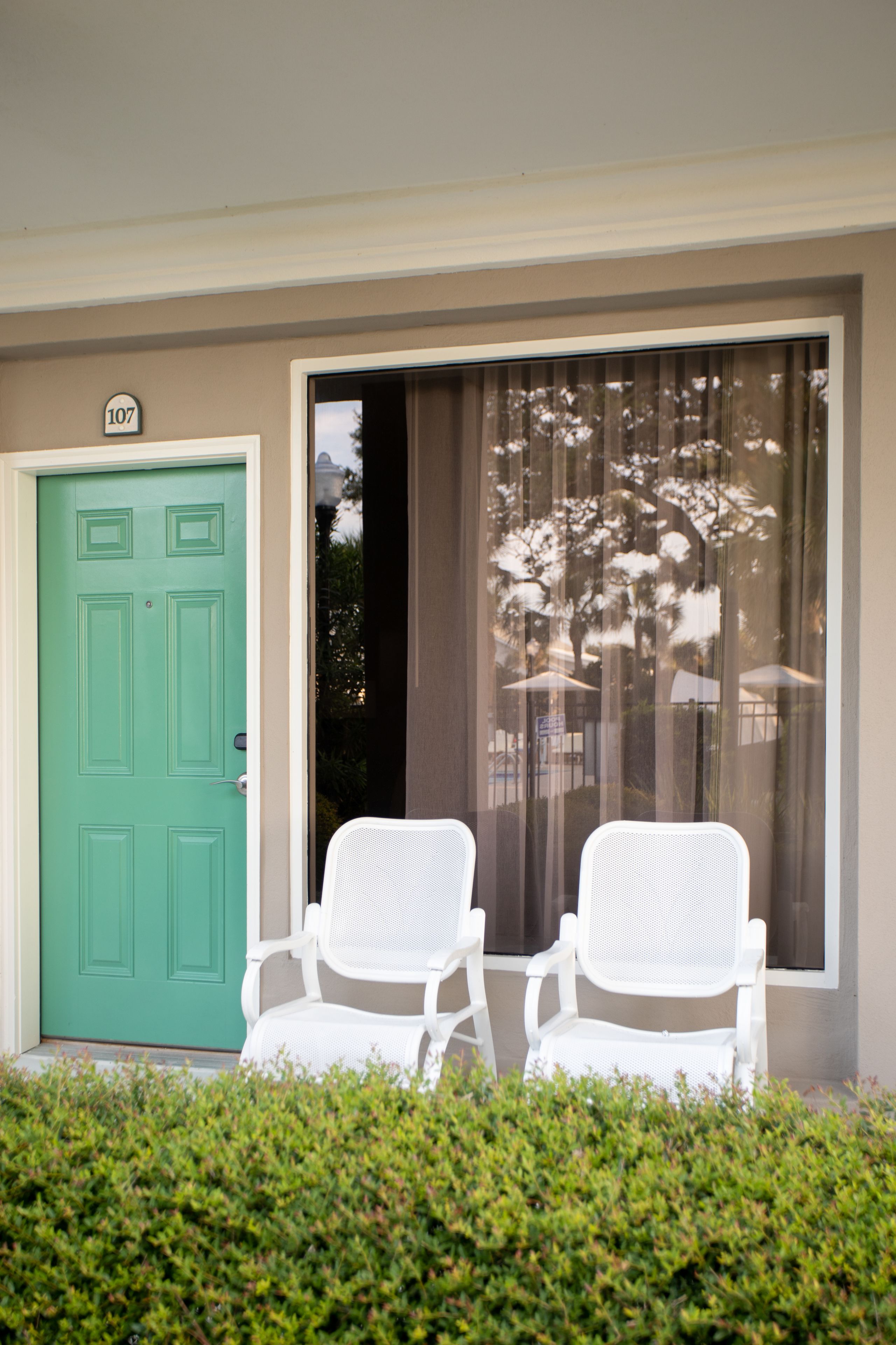 A green door with a number plate, two white chairs in front, and a window with a view of trees and umbrellas.