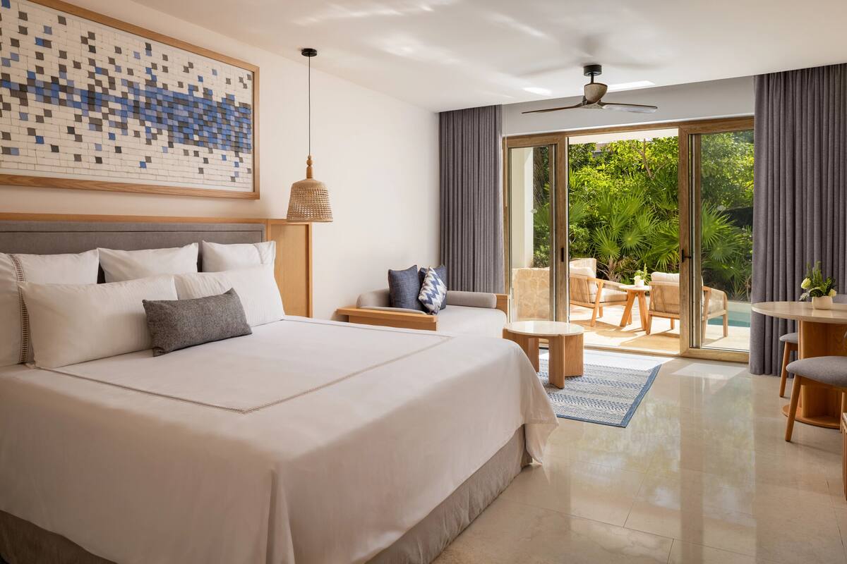 A modern hotel room with a large bed, a ceiling fan, a dining area with a table and chairs, and a sliding glass door to the outside.