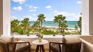 A balcony with wicker chairs, a small table with a vase, and a view of a beach with palm trees and the ocean.