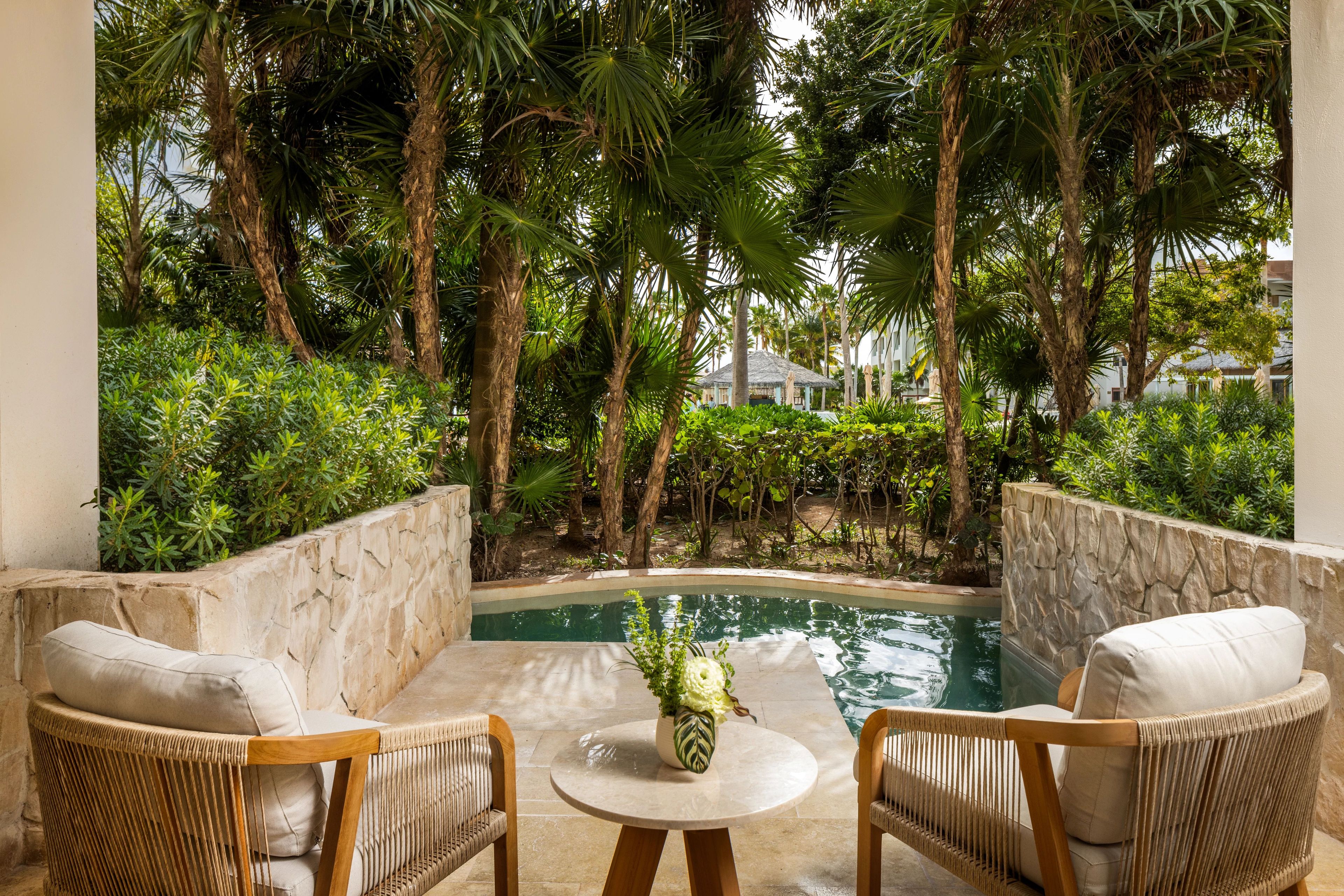 Un área junto a la piscina con dos sillas de mimbre, una pequeña mesa redonda con un florero, y vistas a una piscina rodeada de exuberante vegetación y palmeras.