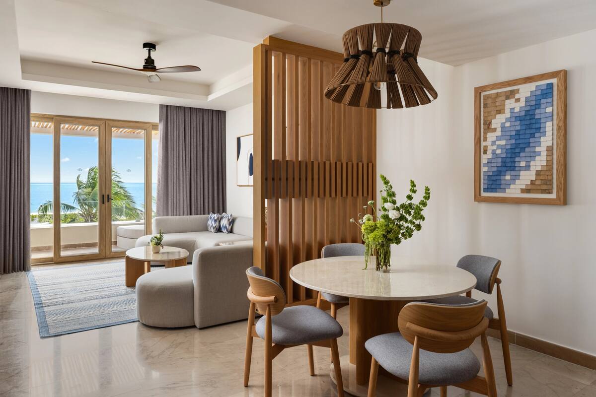 A modern living room with a round dining table, grey upholstered chairs, a ceiling fan, and a view of the ocean.