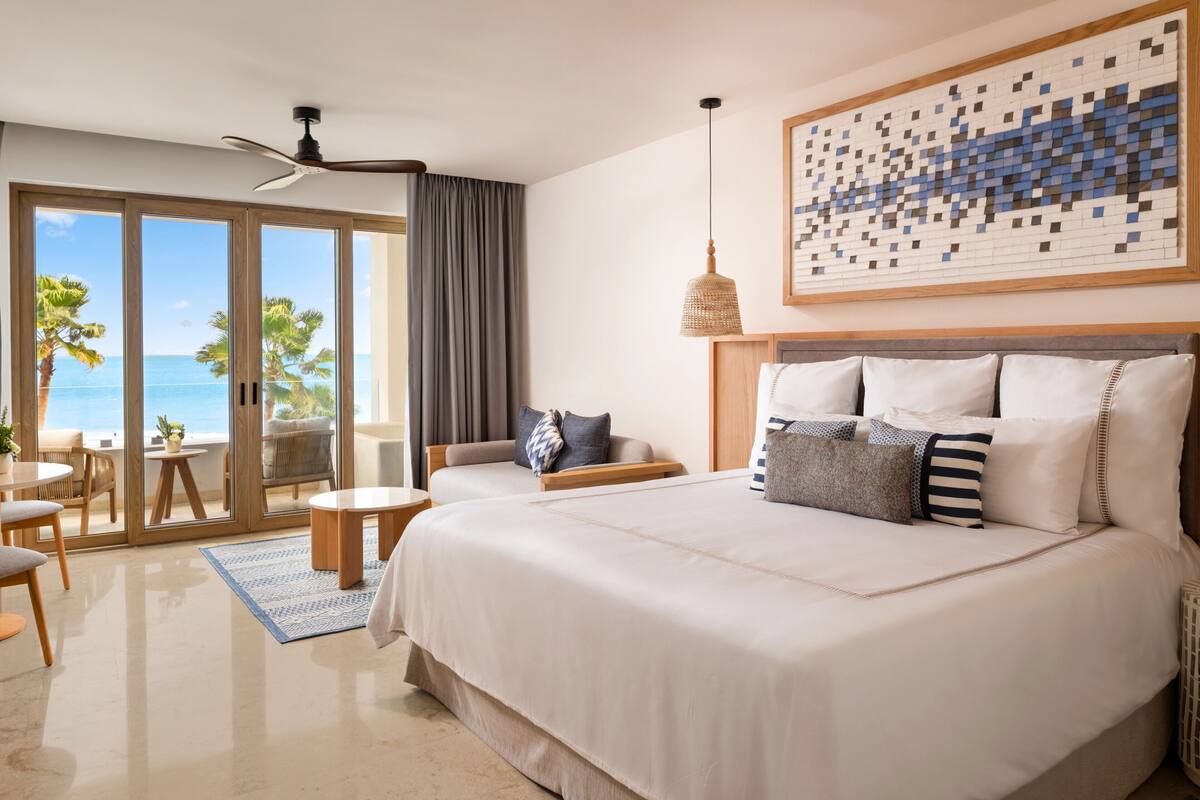 A bedroom with a large bed, a ceiling fan, a view of the ocean through sliding doors, and a mosaic artwork on the wall.