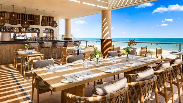 A beachfront restaurant with a long dining table, wicker chairs, and a bar area.