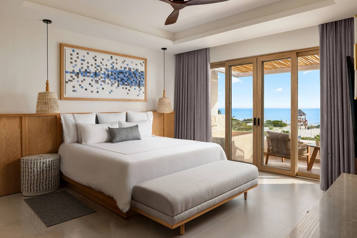 A modern hotel room with a large bed, a view of the ocean, and a balcony with outdoor seating.