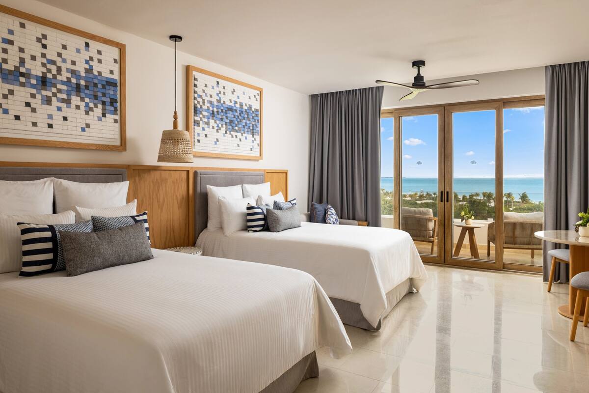 A hotel room with two beds, a balcony with a view of the ocean, and a ceiling fan.