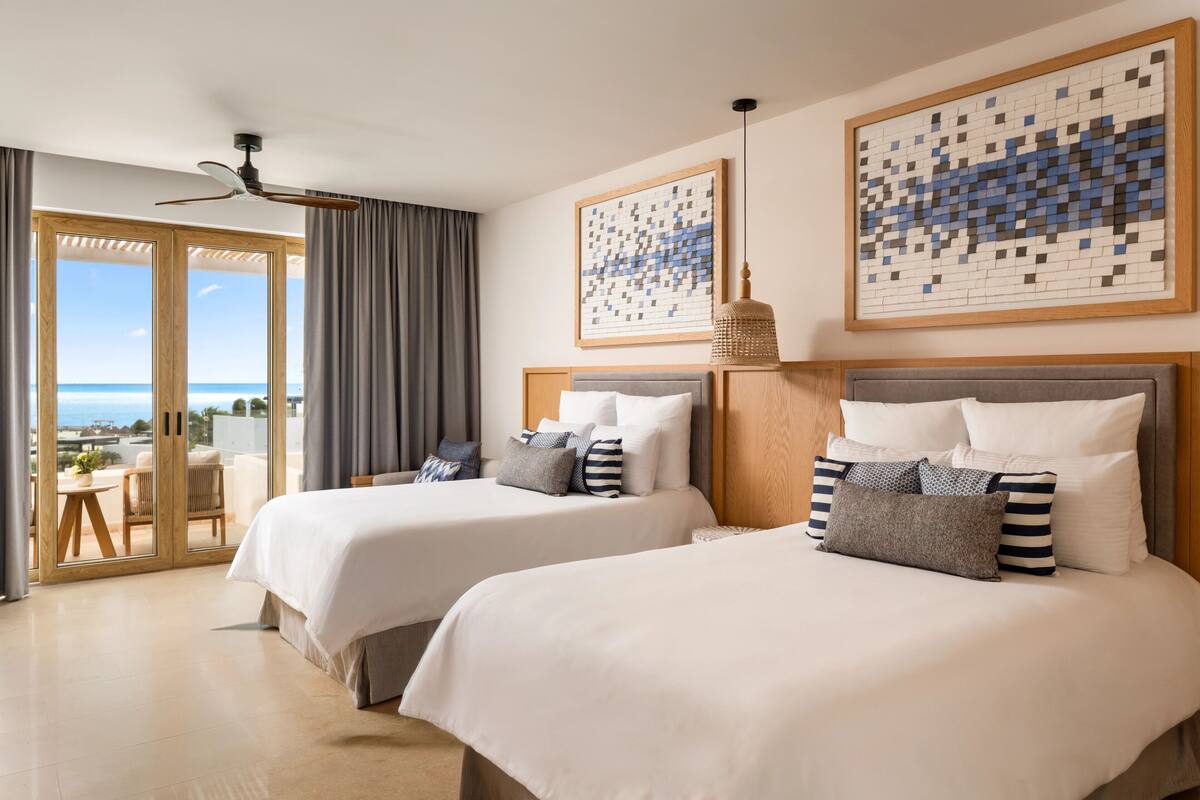 A hotel room with two beds, a ceiling fan, a view through a glass door, and artwork on the walls.
