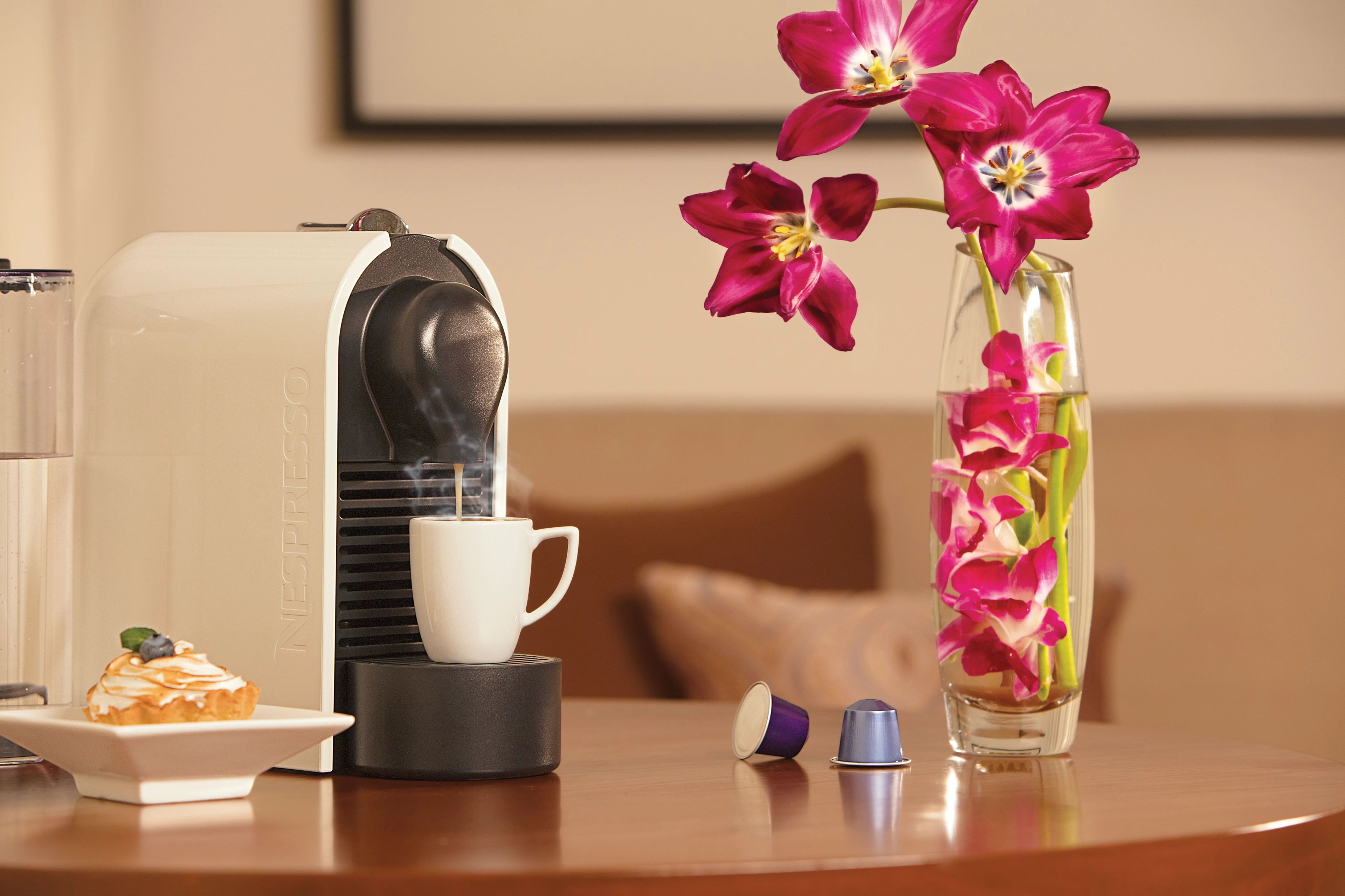 A coffee machine, a white cup, a small plate with a pastry, and a vase with flowers on a table.