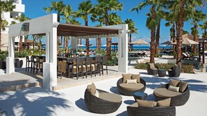 A resort pool area with outdoor seating, a bar, and palm trees.