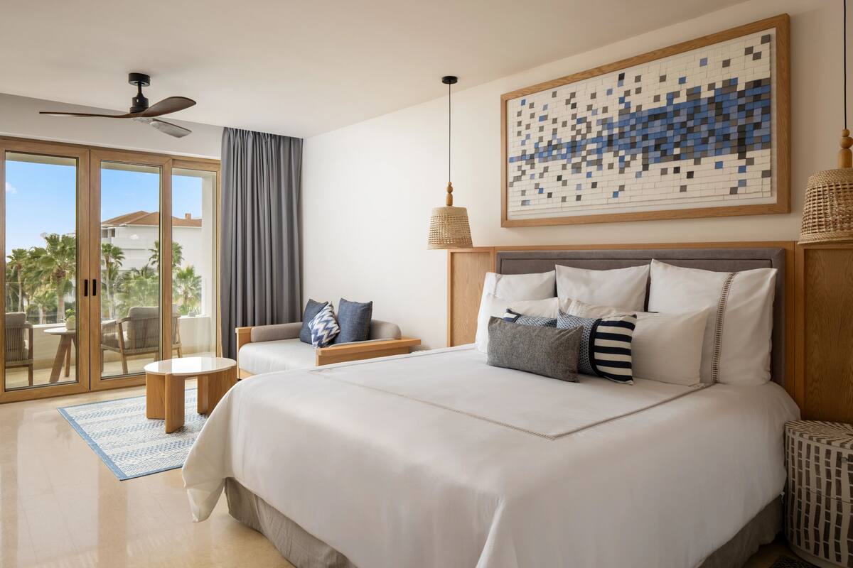 A hotel room with a large bed, a ceiling fan, a view of the outdoors, a small table, and a large abstract painting.