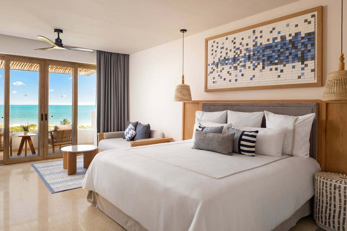 A hotel room with a large bed, a view of the beach, a ceiling fan, and a woven lamp.