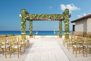 An outdoor wedding venue with a white carpet, golden chairs, and floral arches.
