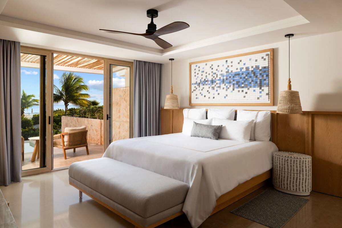 A bedroom with a bed, a ceiling fan, a view of the outdoors, a wall-mounted artwork, and a small table with a chair.