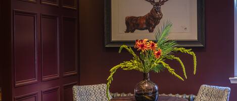 A room with a dark wooden door, a framed deer head on the wall, and a table with a vase of flowers.