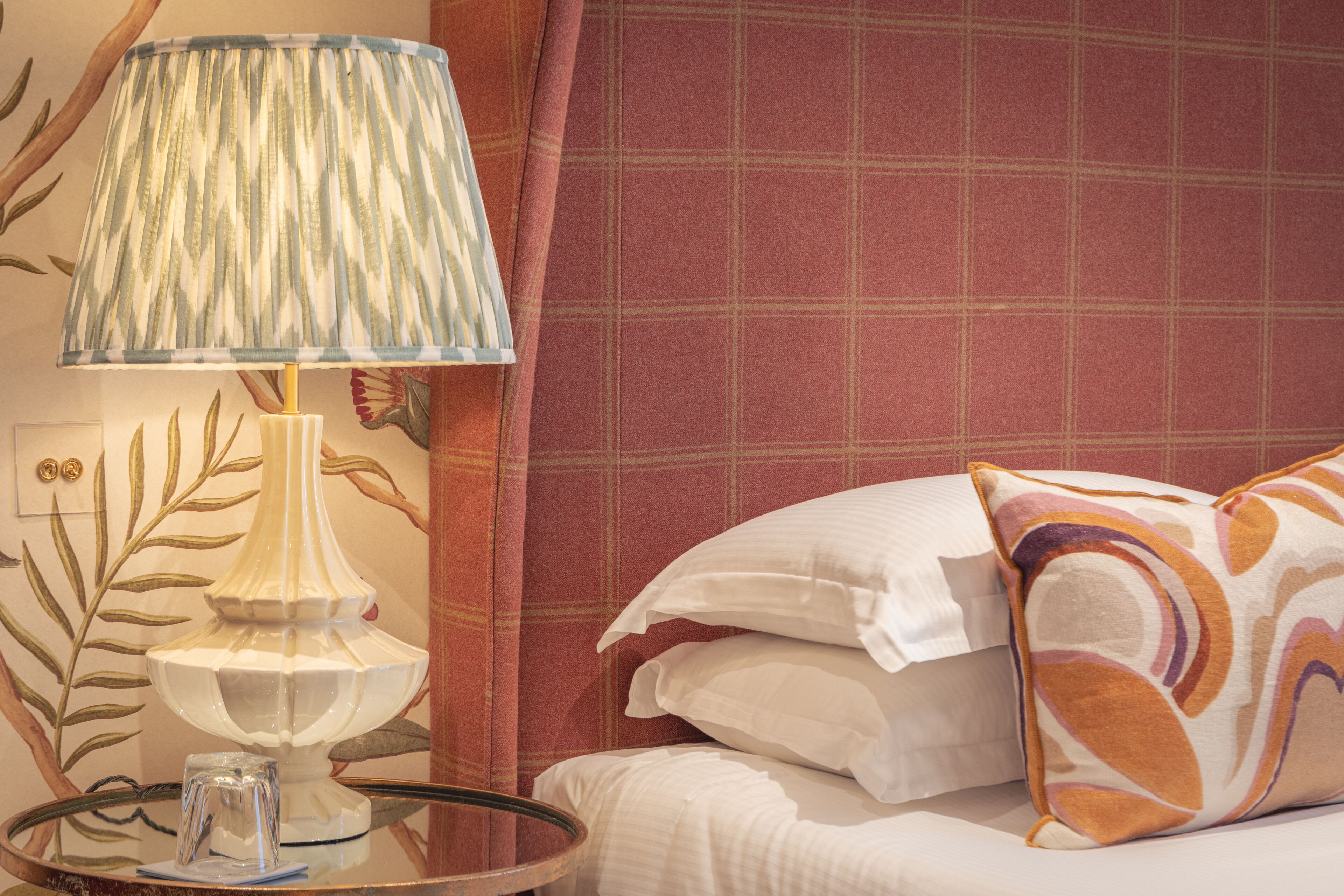 Una habitación de hotel con cama, almohadas, una lámpara y una mesita de vidrio con un florero.