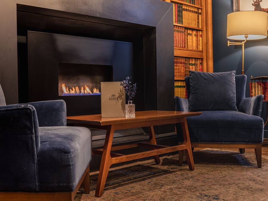 Ein gemütliches Wohnzimmer mit Kamin, zwei blaue Sesseln, einem Holztisch und einem Bücherregal voller Bücher.