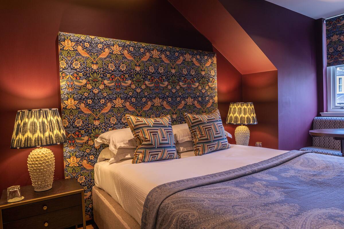 Quarto com cabeceira estampada, duas luminárias de apoio, cama com almofadas padronizadas e janela com cortinas.