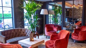 A cozy lobby with a tufted sofa, red velvet chairs, a wooden coffee table, and a large potted plant. - The Raeburn (Edinburgh)