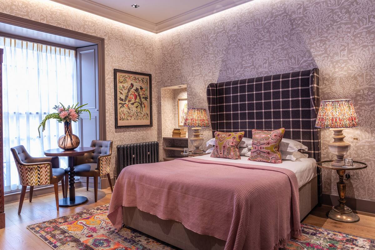 Quarto com uma cama grande, duas mesas de cabeceira, uma cadeira, uma mesinha com um vaso de flores e uma janela com cortinas.