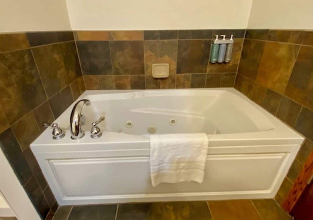 A white freestanding bathtub with a towel, set against a tiled wall with a mix of brown and beige tiles.