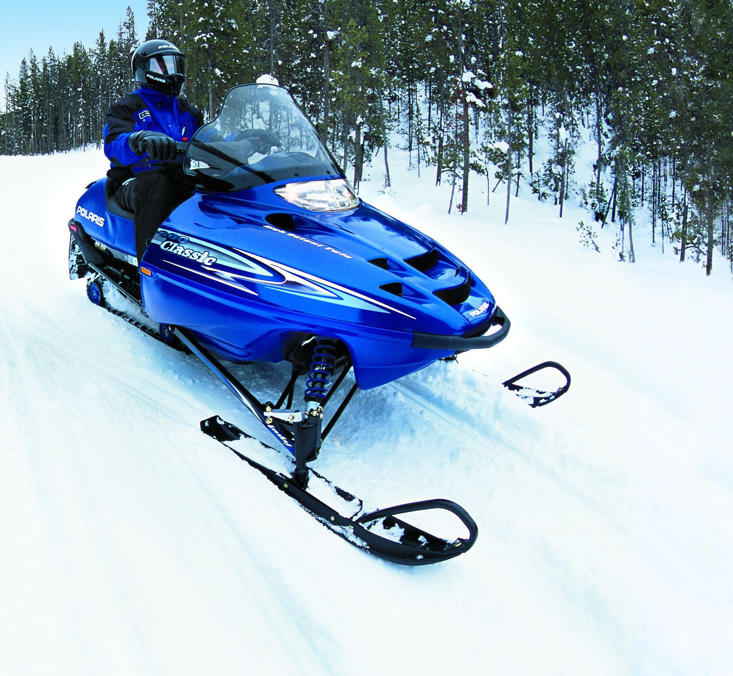 Une motoneige Polaris bleue, avec un conducteur vêtu d’une combinaison bleue, emprunte un sentier enneigé à travers une forêt.