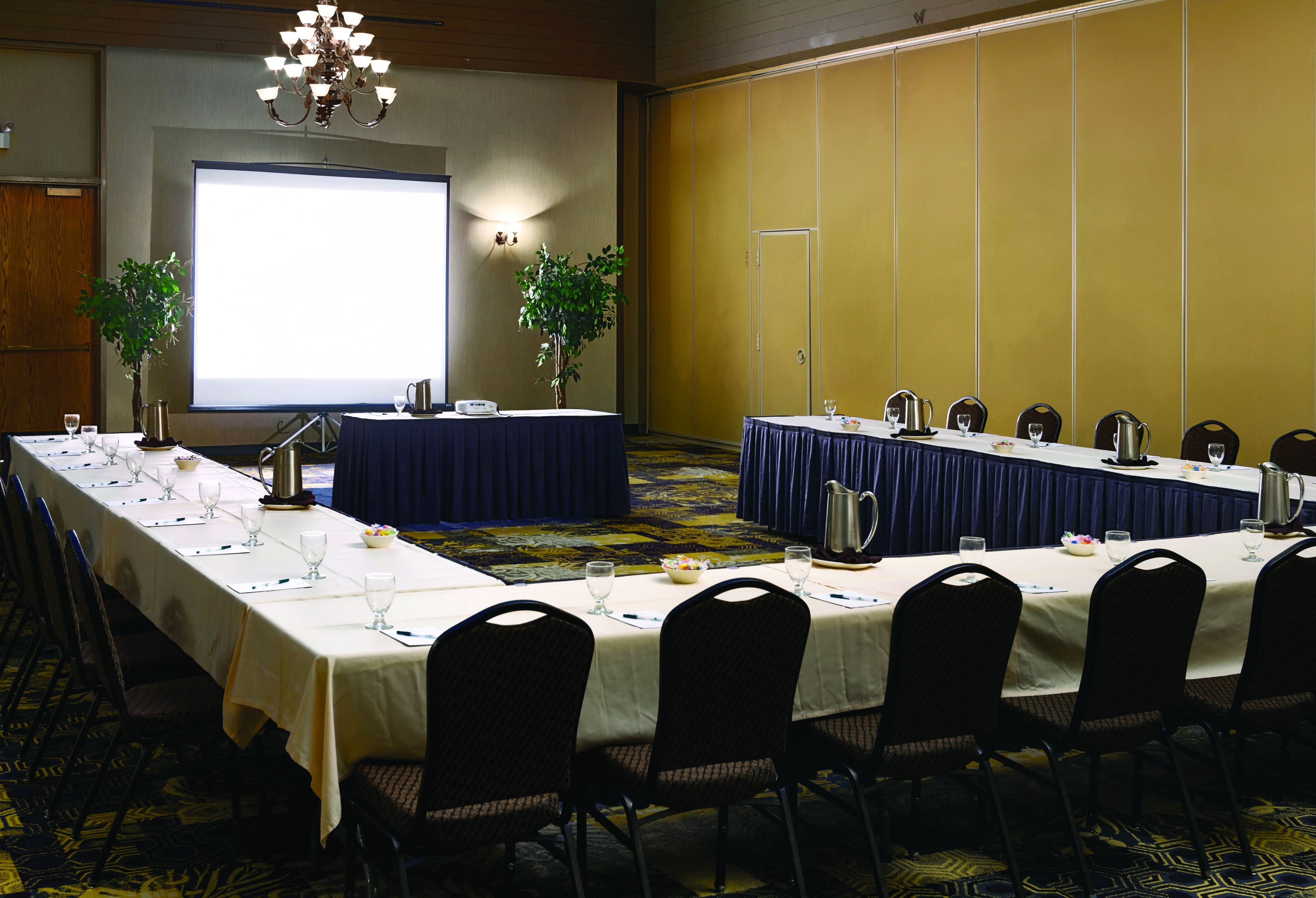 A conference room with long tables, chairs, a projector screen, and two potted plants.