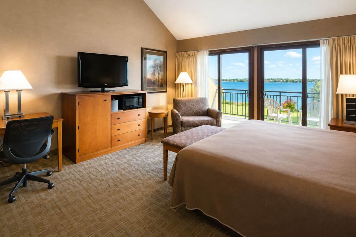 A hotel room with a bed, a desk with a computer, a TV, a chair, and a view of the water.